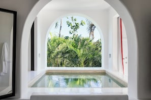 Une pièce avec vue sur une végétation luxuriante, grâce à une fenêtre en demi-cercle, une petite piscine et des murs blancs.