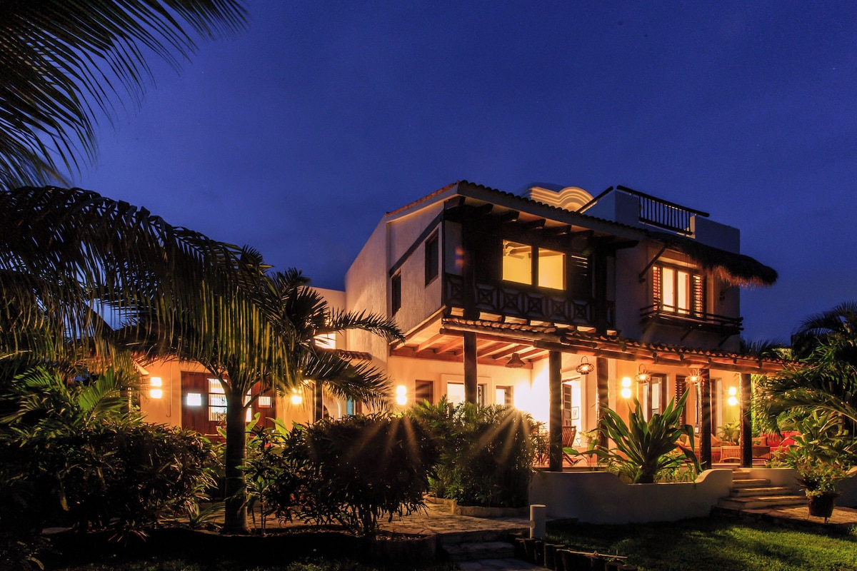 Una casa de dos pisos con un balcón, rodeada de vegetación tropical.