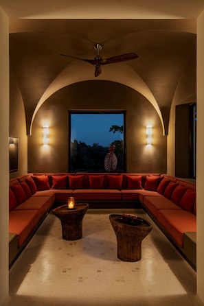 A modern interior with a curved ceiling, a large window with a view of trees, a red sectional sofa, and two wooden coffee tables with candles.