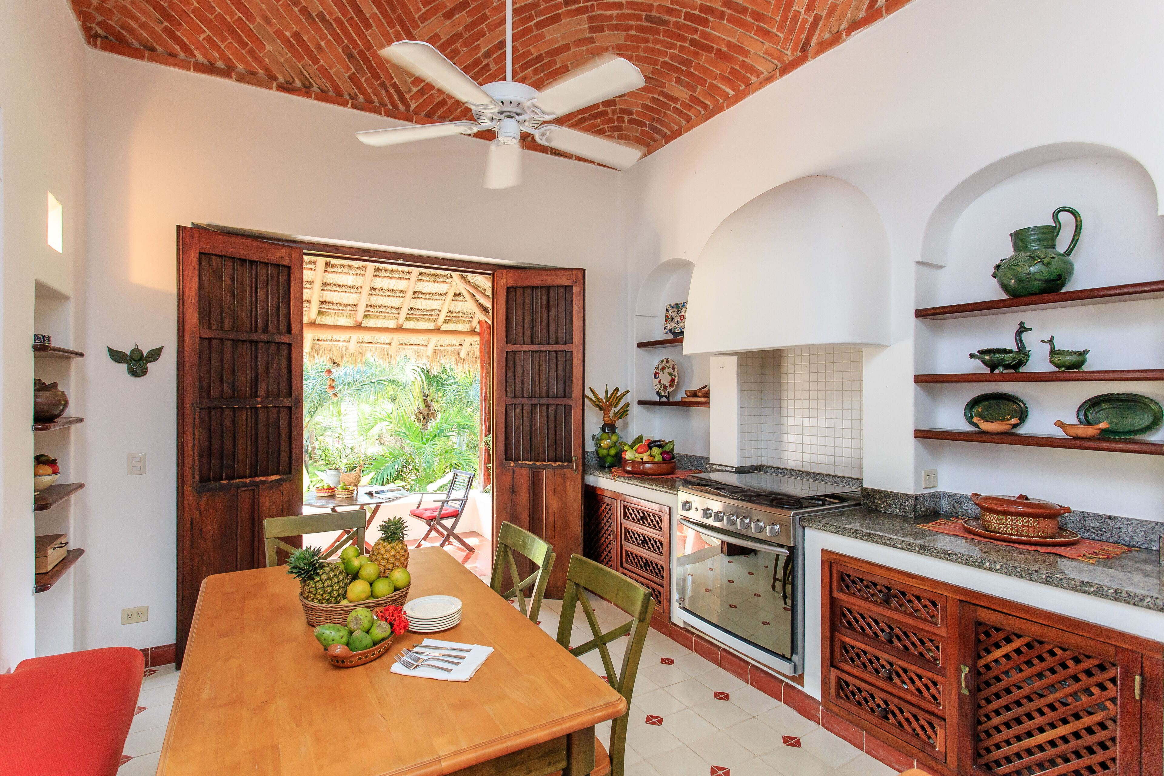 A kitchen with a wooden table, chairs, and a ceiling fan. There is a window with a view of greenery outside.