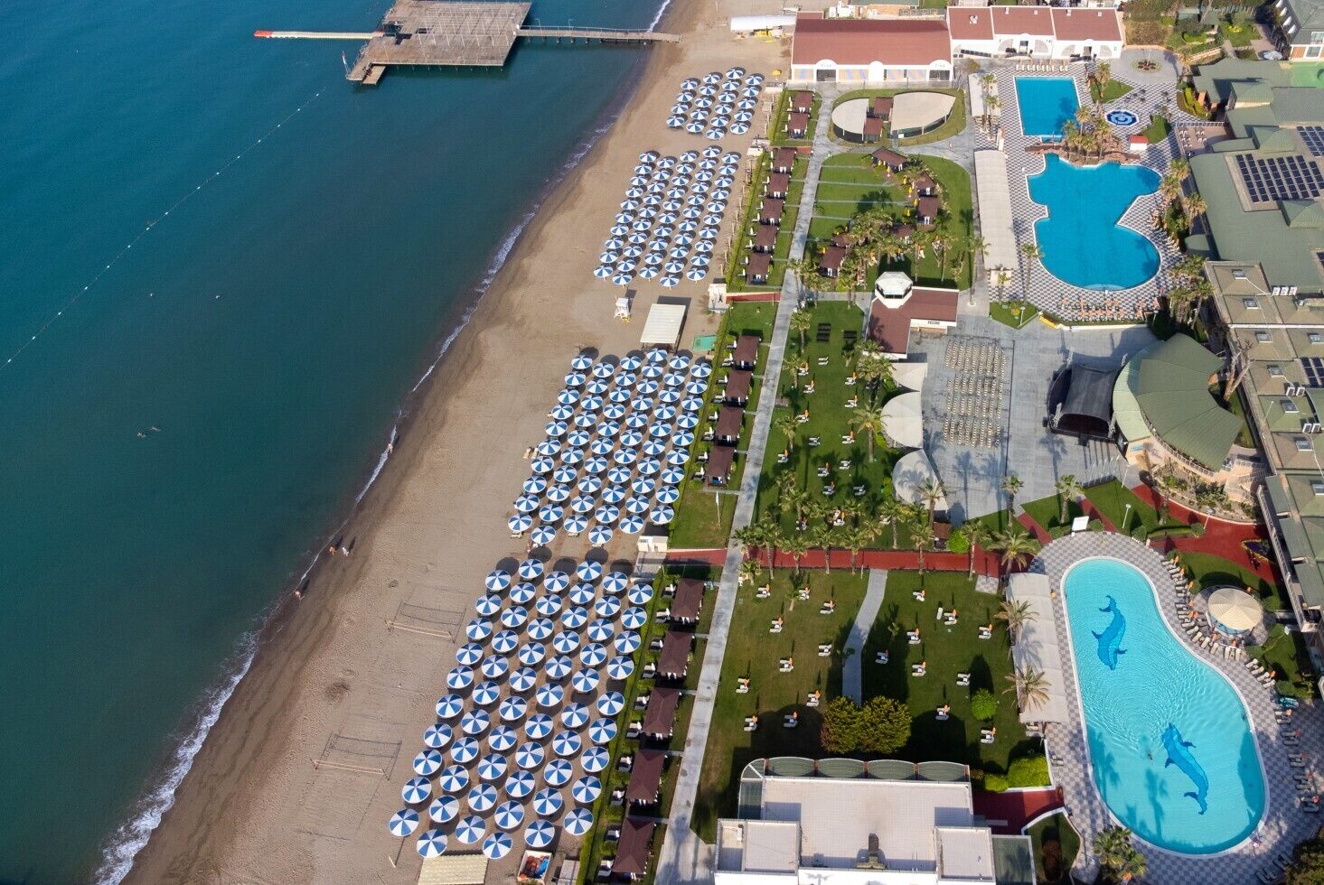 An aerial view of a resort with a beach, multiple swimming pools, and numerous sun umbrellas.