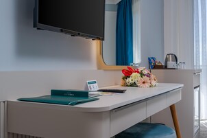 Habitación de hotel moderna con un escritorio blanco, un televisor de pantalla plana montado encima, un pequeño jarrón con flores, una cafetera y una ventana con cortinas.