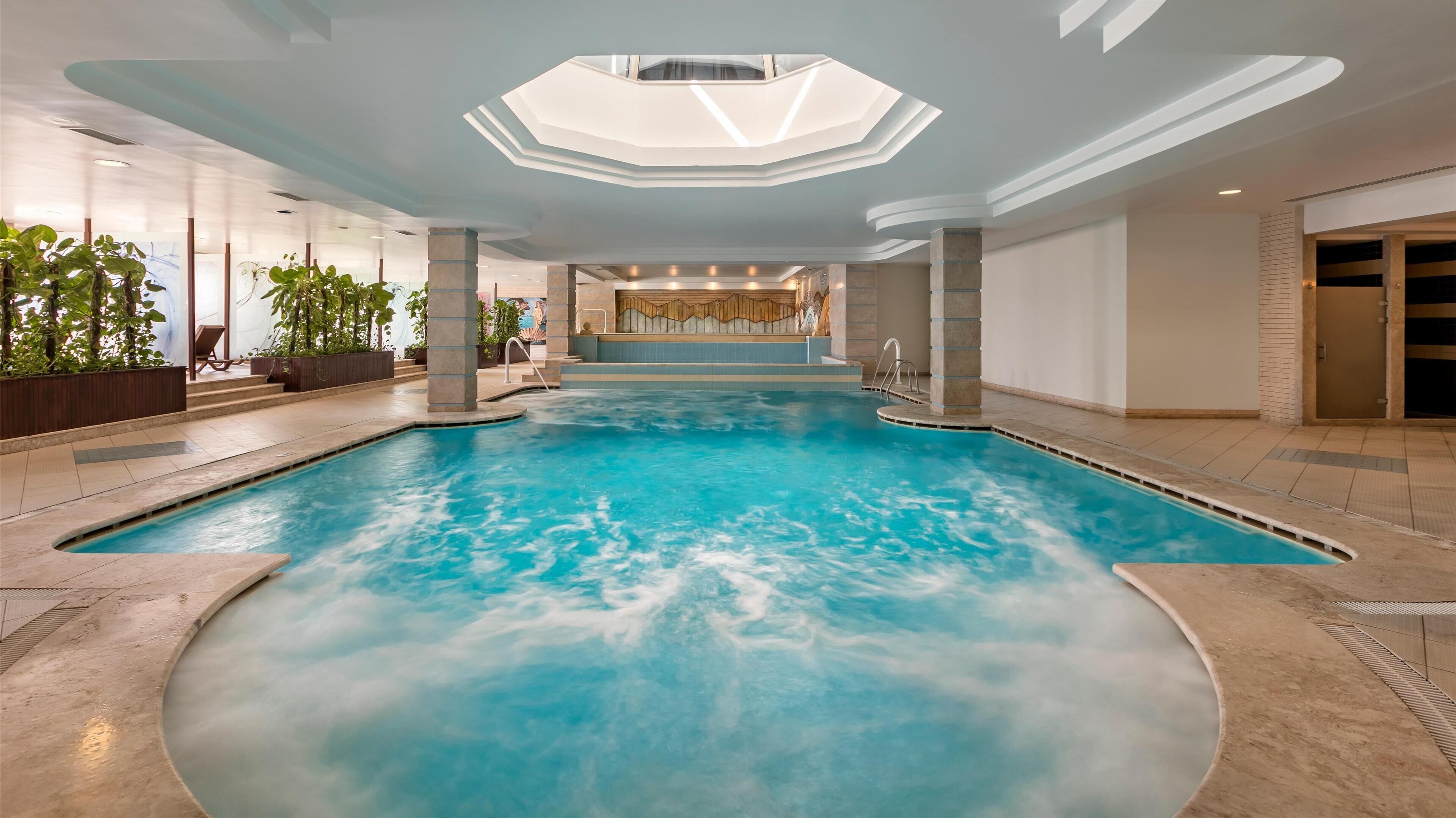 Una amplia piscina interior con techo de tragaluces, rodeada de paredes de piedra y plantas en macetas.