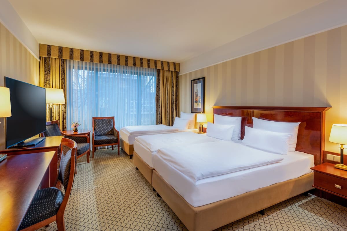 Ein Hotelzimmer mit zwei Betten, einem Schreibtisch mit Fernseher, einem Sessel und einem Fenster mit Vorhängen.