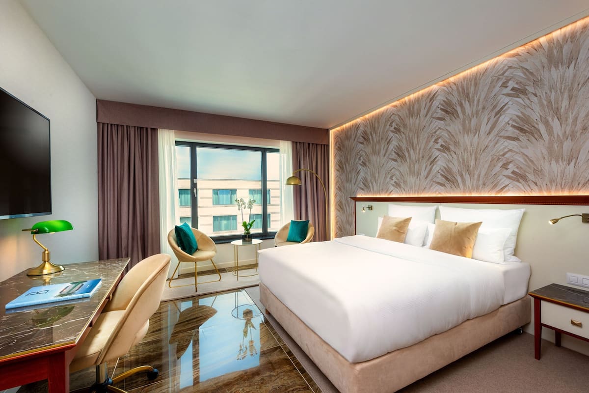 Ein Hotelzimmer mit einem großen Bett, einem Schreibtisch mit Lampe, zwei Stühlen, einem Fernseher und Blick auf die Stadt durch das Fenster.