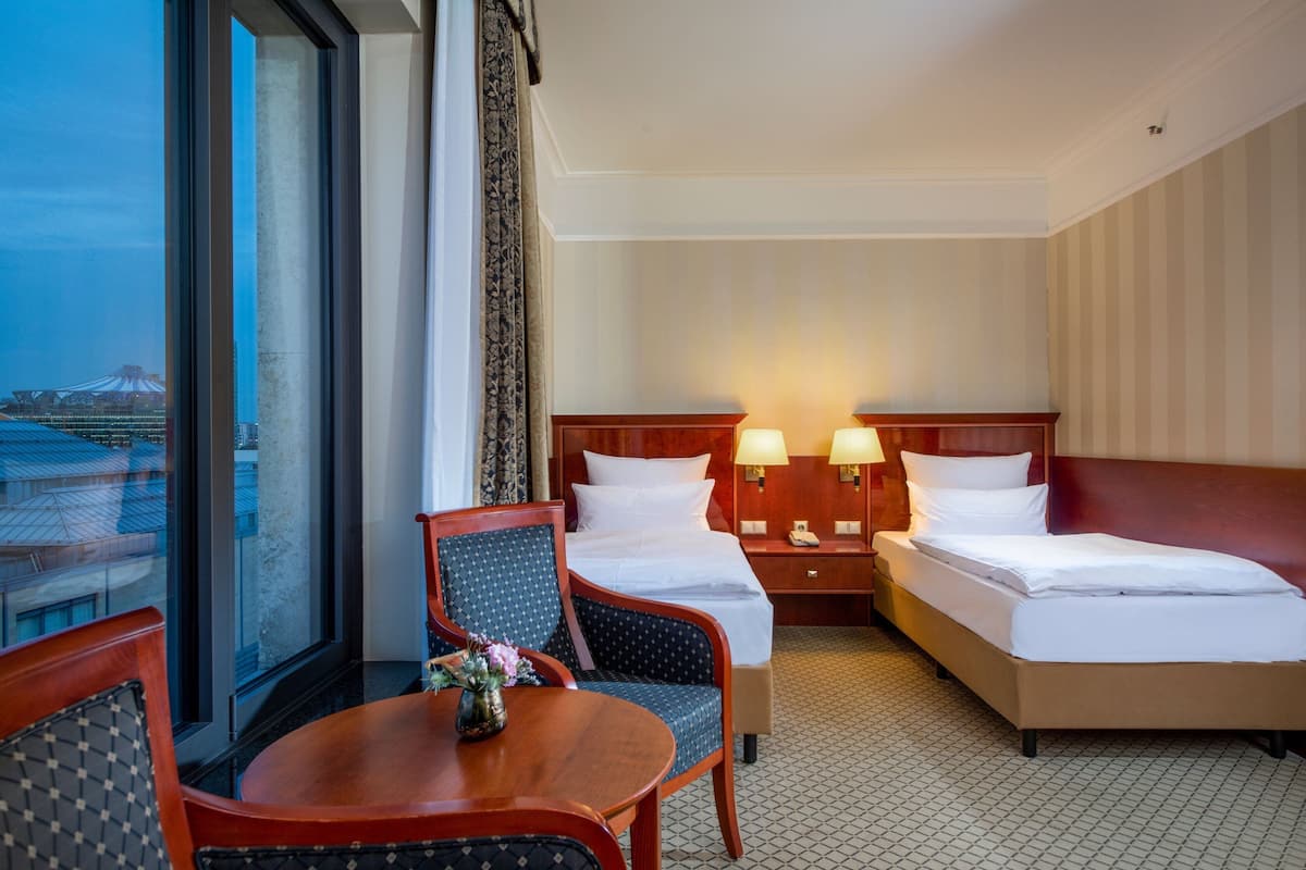Ein Hotelzimmer mit zwei Betten, einem kleinen Tisch mit einer Vase, einem Sessel und Blick auf eine Stadtlandschaft durch große Fenster.