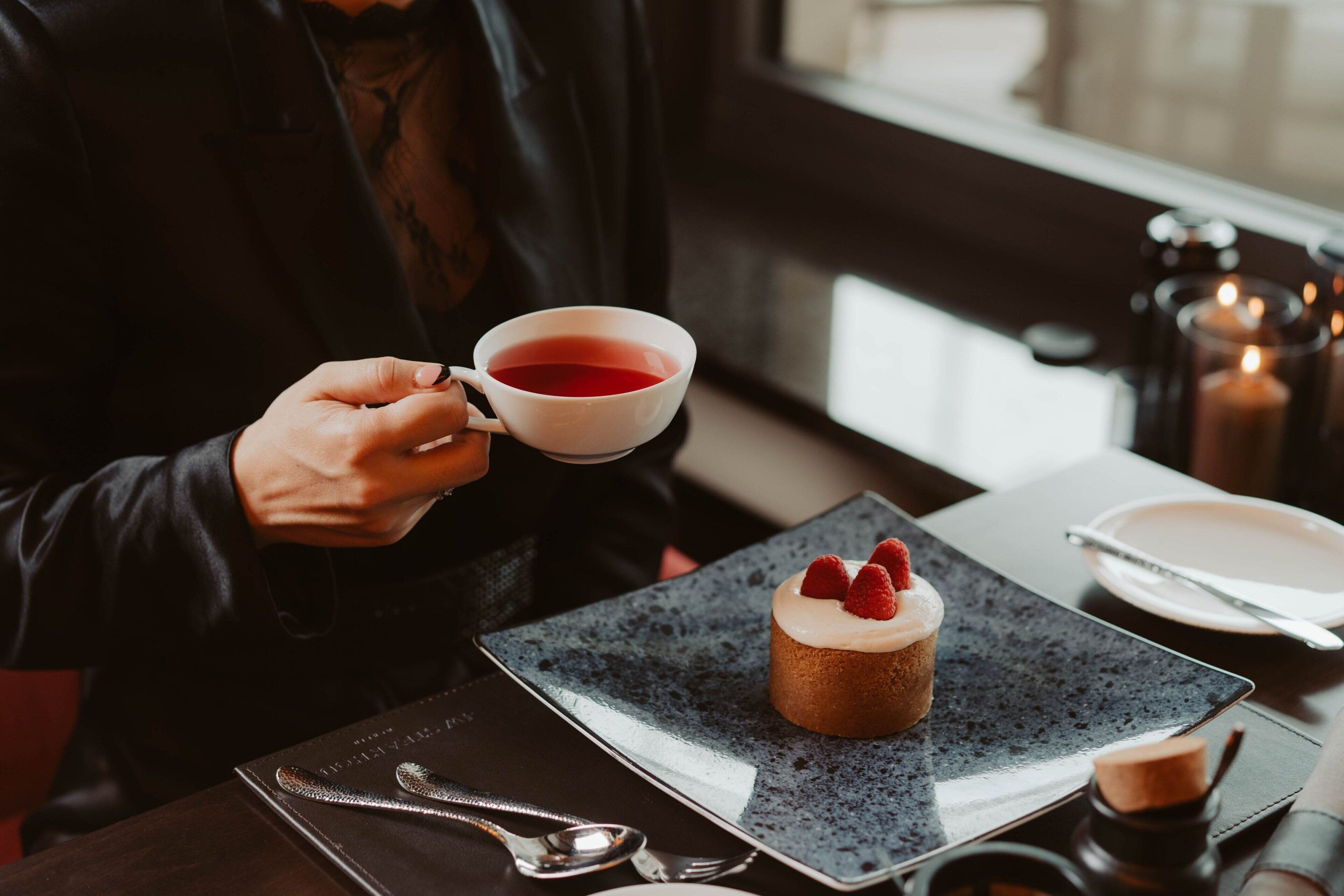 Iemand houdt een kop thee vast, een dessert met aardbeien, en een bord met een lepel op een tafel.
