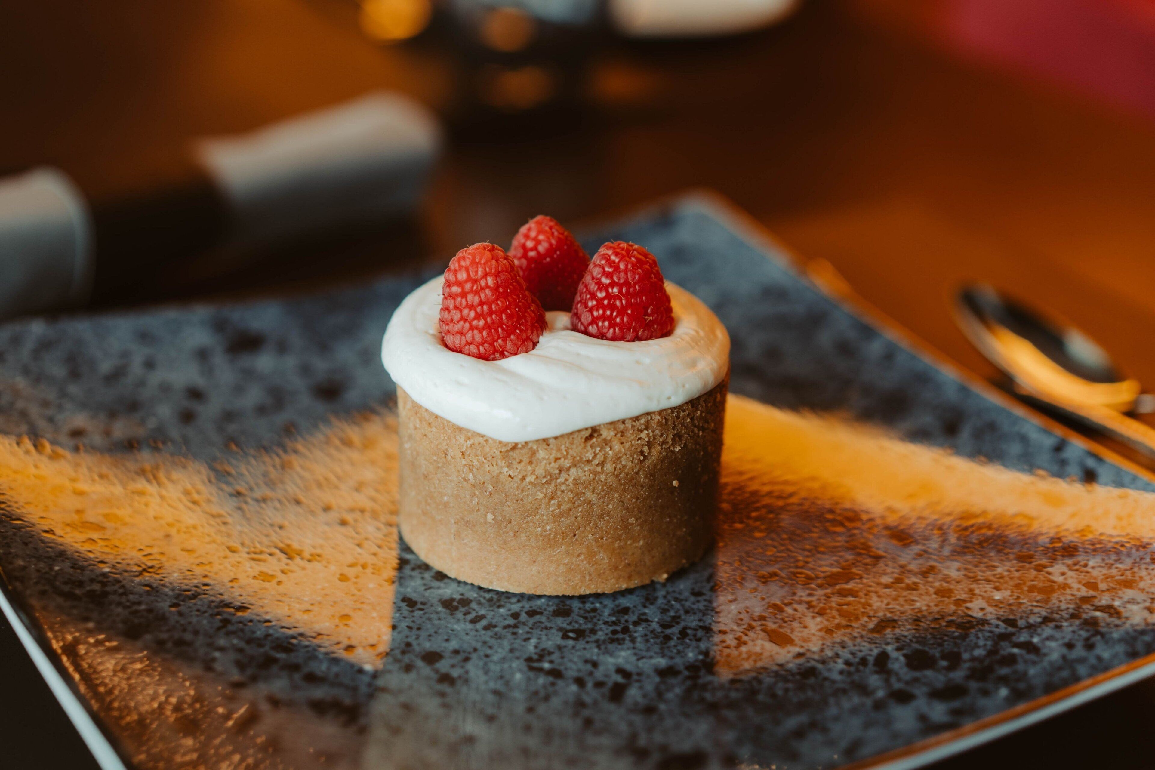 A dessert with three raspberries on top, placed on a square plate with a textured surface.