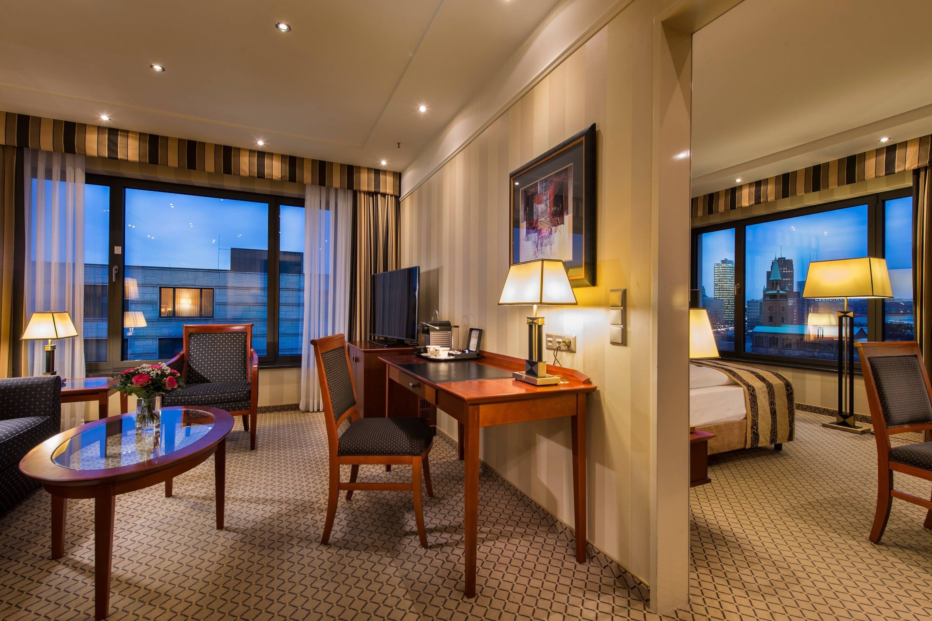 A hotel room with a bed, a desk, two chairs, a TV, and large windows with a city view.