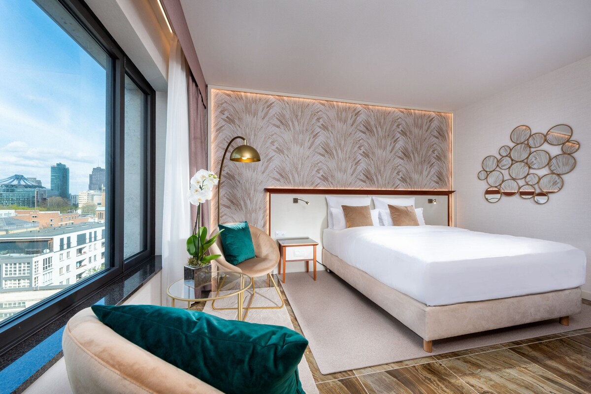 Un hotel moderno con una cama grande, una silla, una mesita de noche con una lámpara y vistas de la ciudad a través de amplios ventanales.