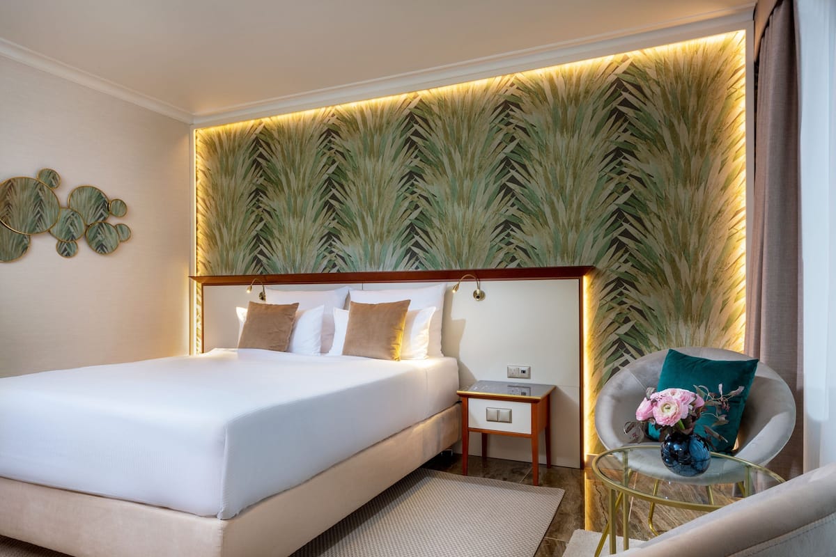 Ein modernes Hotelzimmer mit einem großen Bett, einem Nachttisch, einem Sessel mit Blumen in einer Vase und einer Wand mit einem Blattmuster.