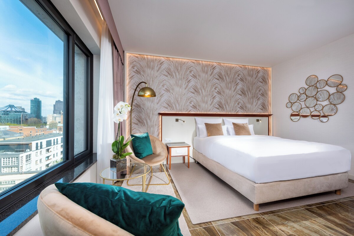 Una habitación de hotel moderna con una cama grande, un área de descanso con una silla y una mesa, vistas a la ciudad y un panel decorativo en la pared.