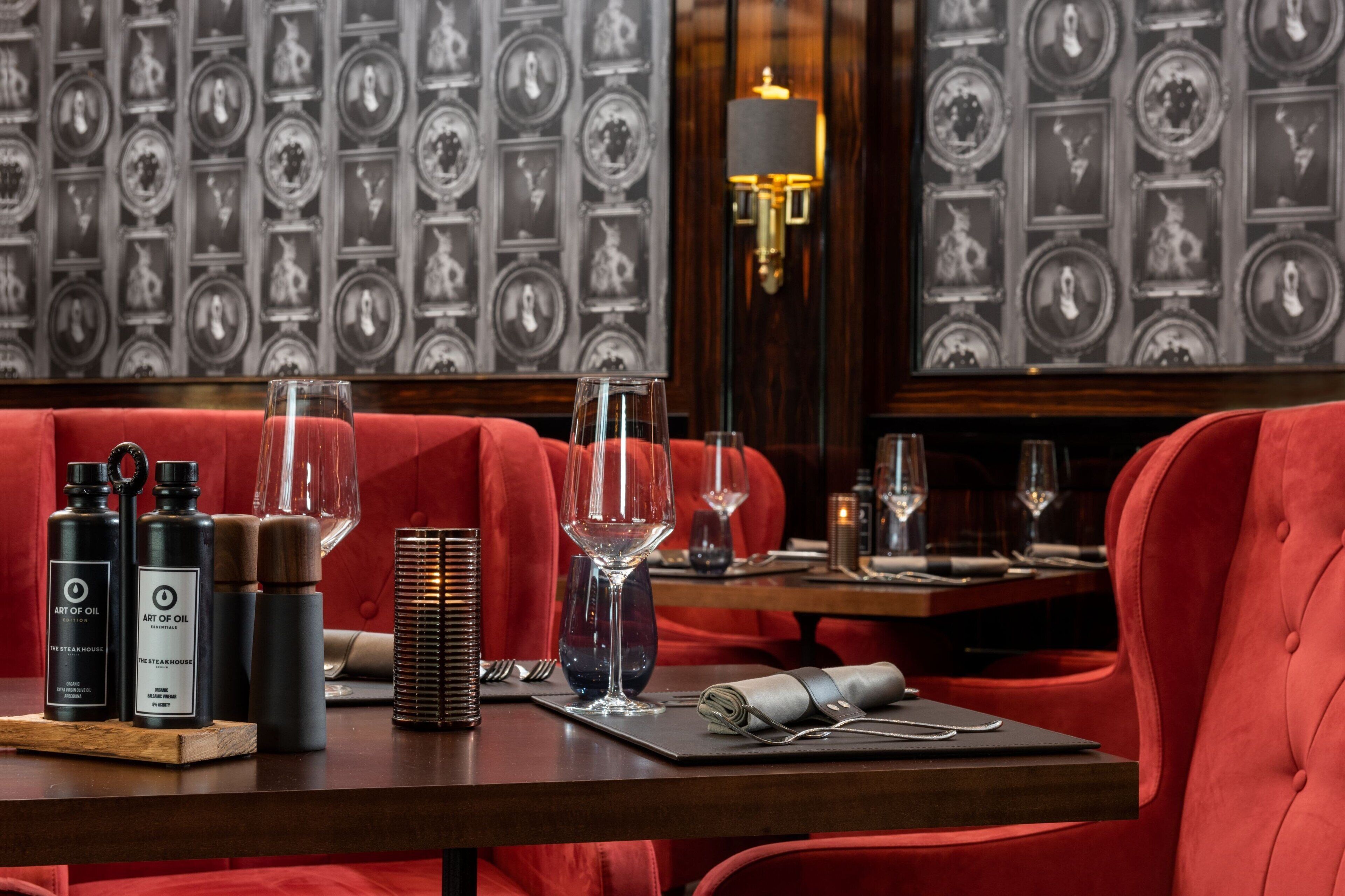 Interior de restaurante con asientos tapizados en rojo, una mesa de madera con copas y botellas de vino, y una pared con retratos enmarcados.