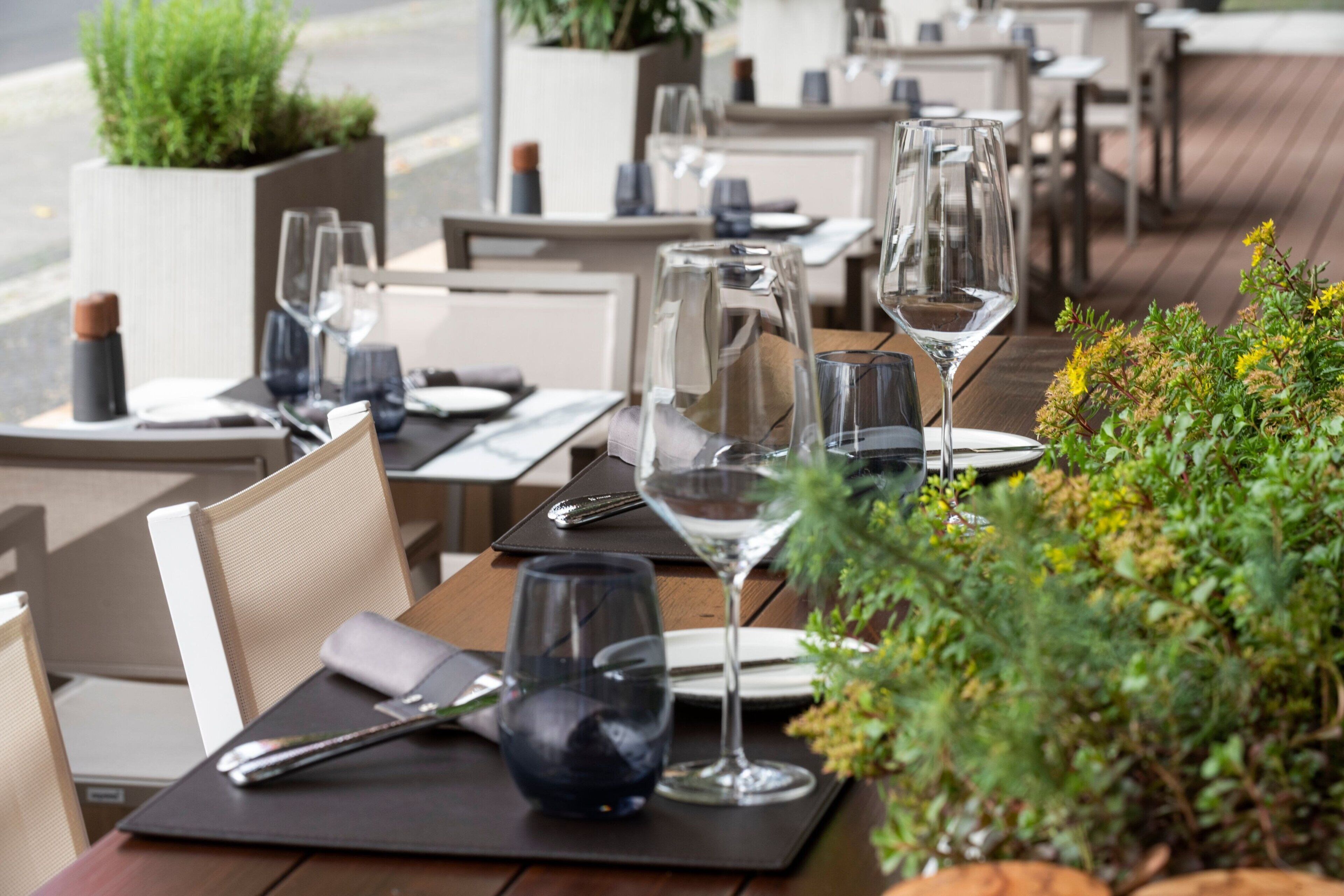 Un restaurante con terraza, mesas ordenadas con copas de vino y cubiertos, y plantas en macetas que aportan verdor.