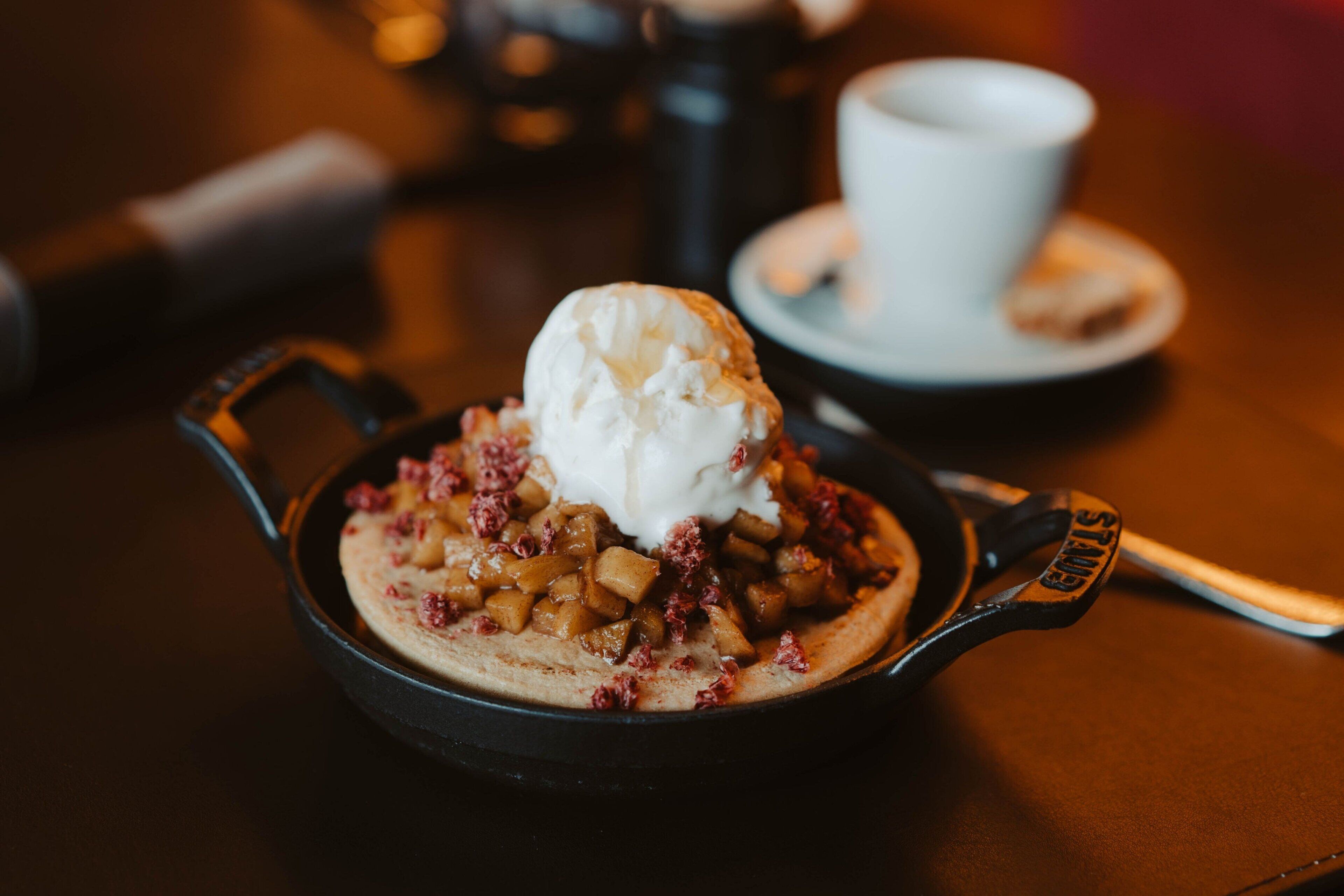 A skillet with a dessert topped with ice cream and diced fruit, accompanied by a cup and saucer on a table.