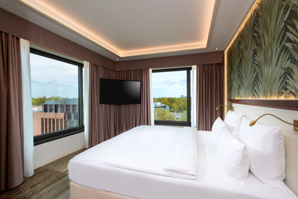 Habitación de hotel con una cama grande, un televisor de pantalla plana, un ventanal con cortinas y vistas a edificios y árboles.