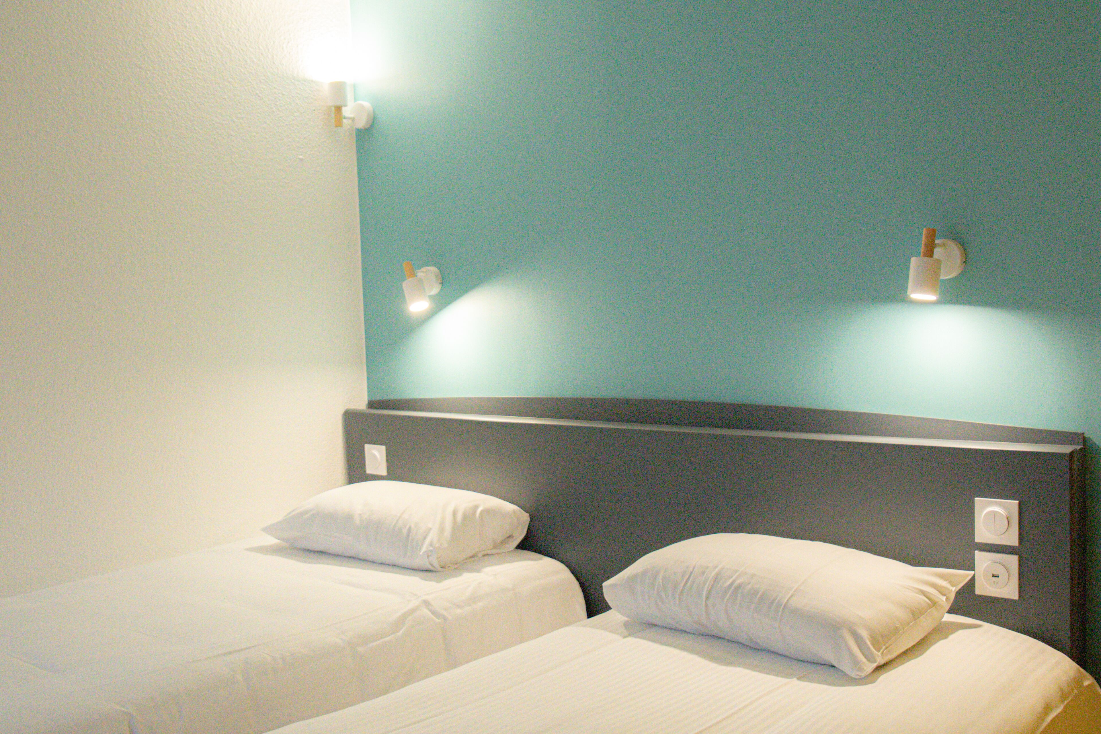 Habitación de hotel con dos camas individuales, cabecera oscura y lámparas fijadas a la pared.