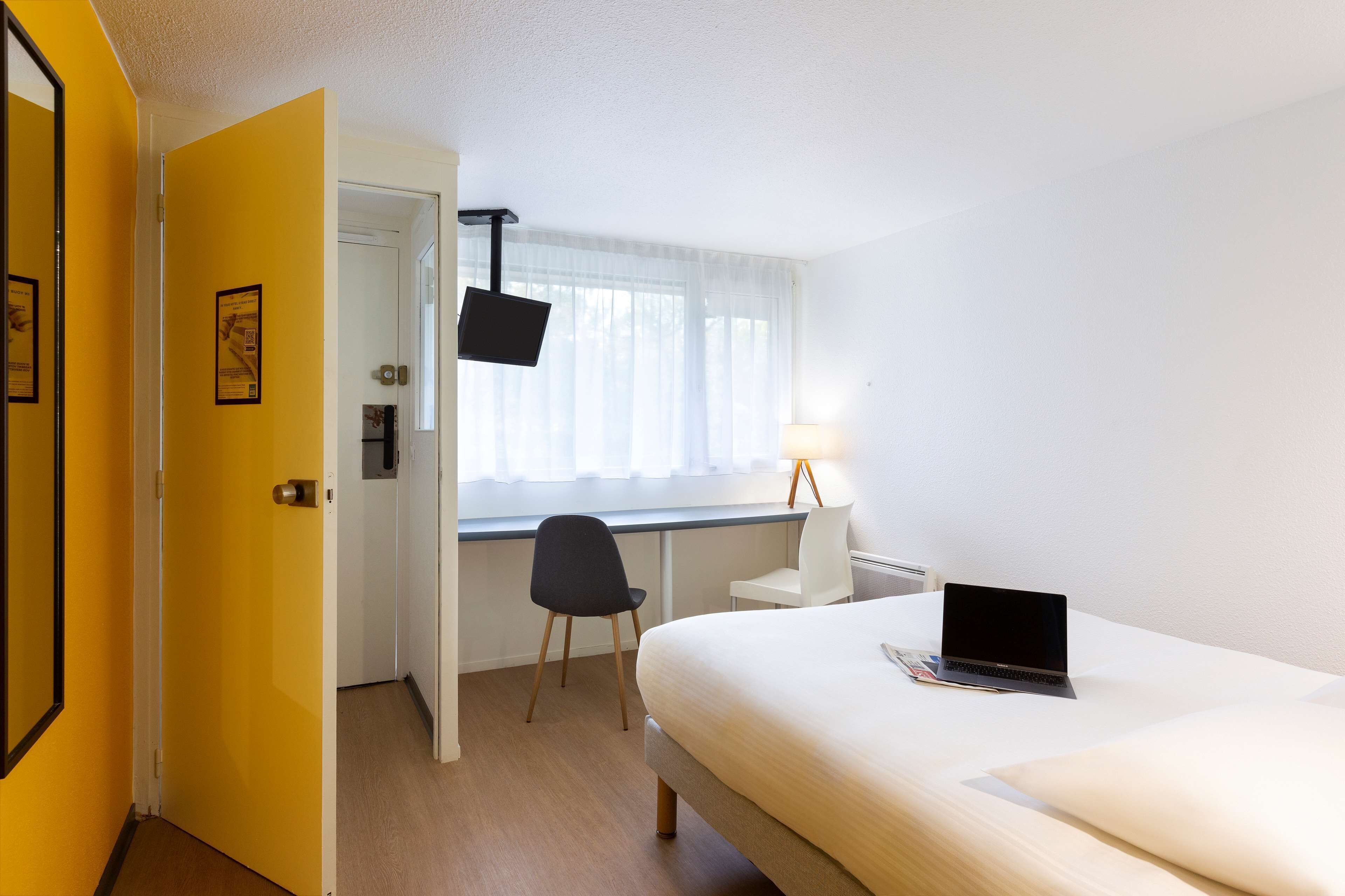 A hotel room with a bed, a desk with a laptop, a chair, a window with curtains, and a door.