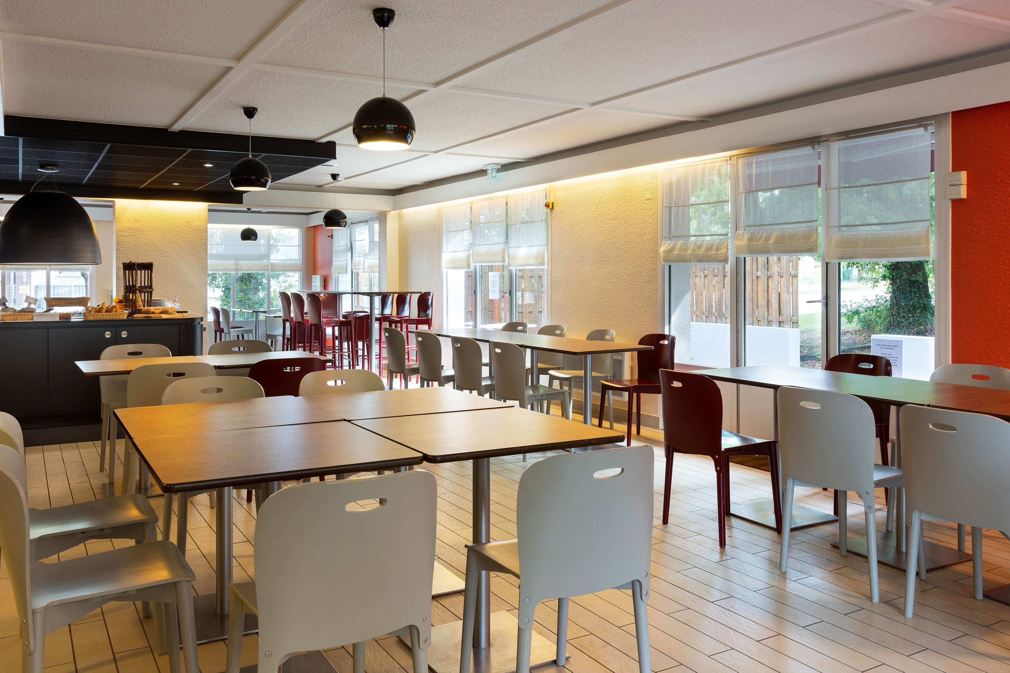 A modern dining area with multiple tables and chairs, large windows with blinds, and a kitchen area in the background.