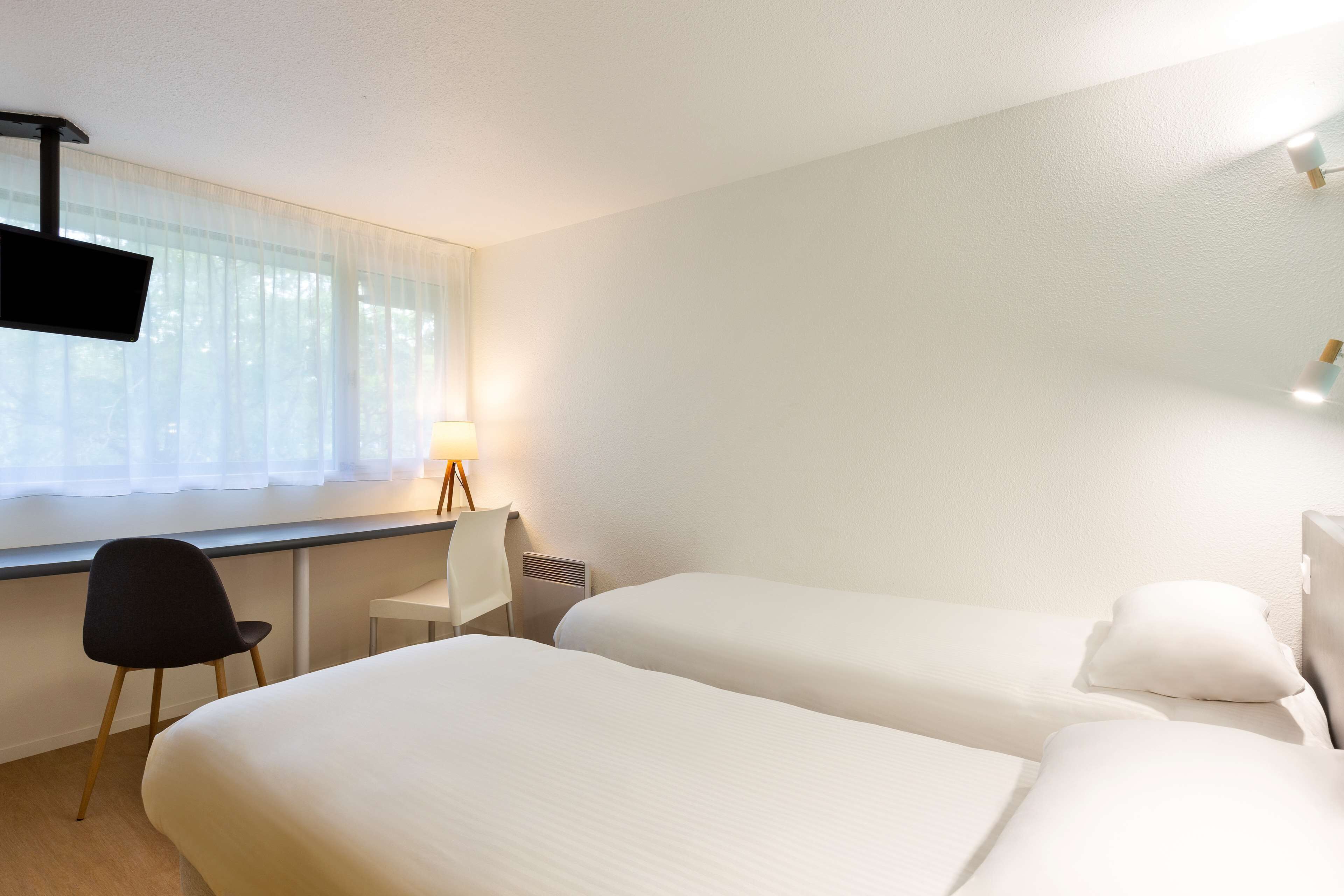 Ein Hotelzimmer mit zwei Betten, einem Schreibtisch, einem Stuhl, einem Fernseher und einem Fenster mit leichtem Vorhang.