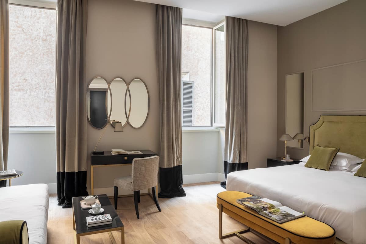 A hotel room with a bed, a desk with a chair, a small table with a book and a teapot, and a window with curtains.