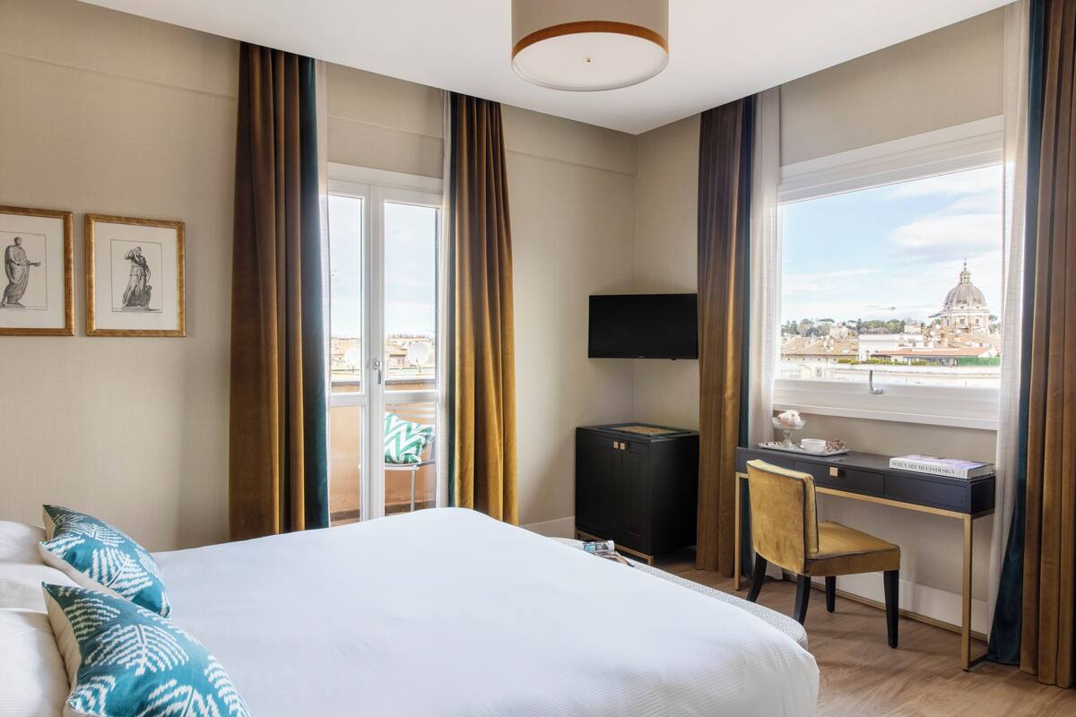 A hotel room with a bed, a desk with a chair, a TV, and a window with a view of a dome and buildings.