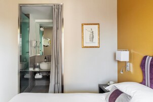 A hotel room with a bed, a nightstand, a wall-mounted lamp, a framed artwork, and a bathroom visible through an open door.