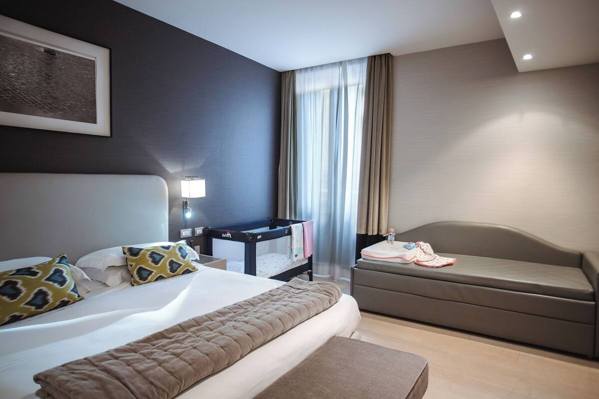 A modern hotel room with a large bed, a sofa, a desk, and a window with curtains.