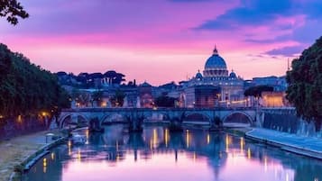 A bridge over a calm river reflecting a historic building with a dome, surrounded by trees and city lights.