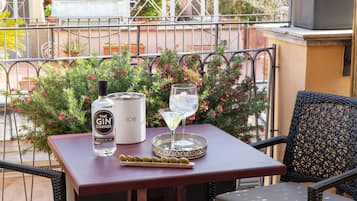 Una mesa con una botella de ginebra, un vaso de agua y un plato de aceitunas en un balcón con barandilla y plantas en macetas.