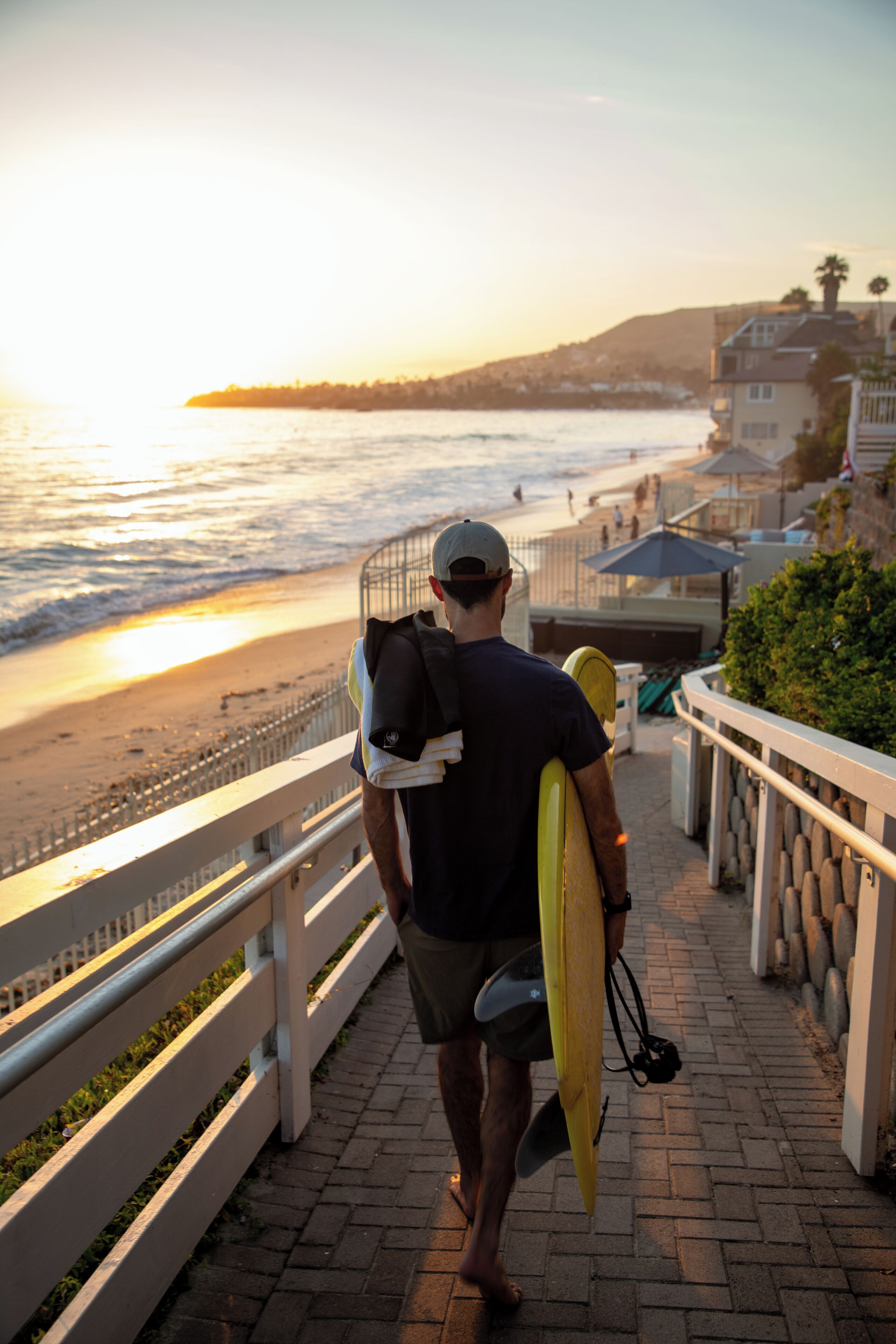 A man walking on a paved path with a surfboard, overlooking a beach and ocean.