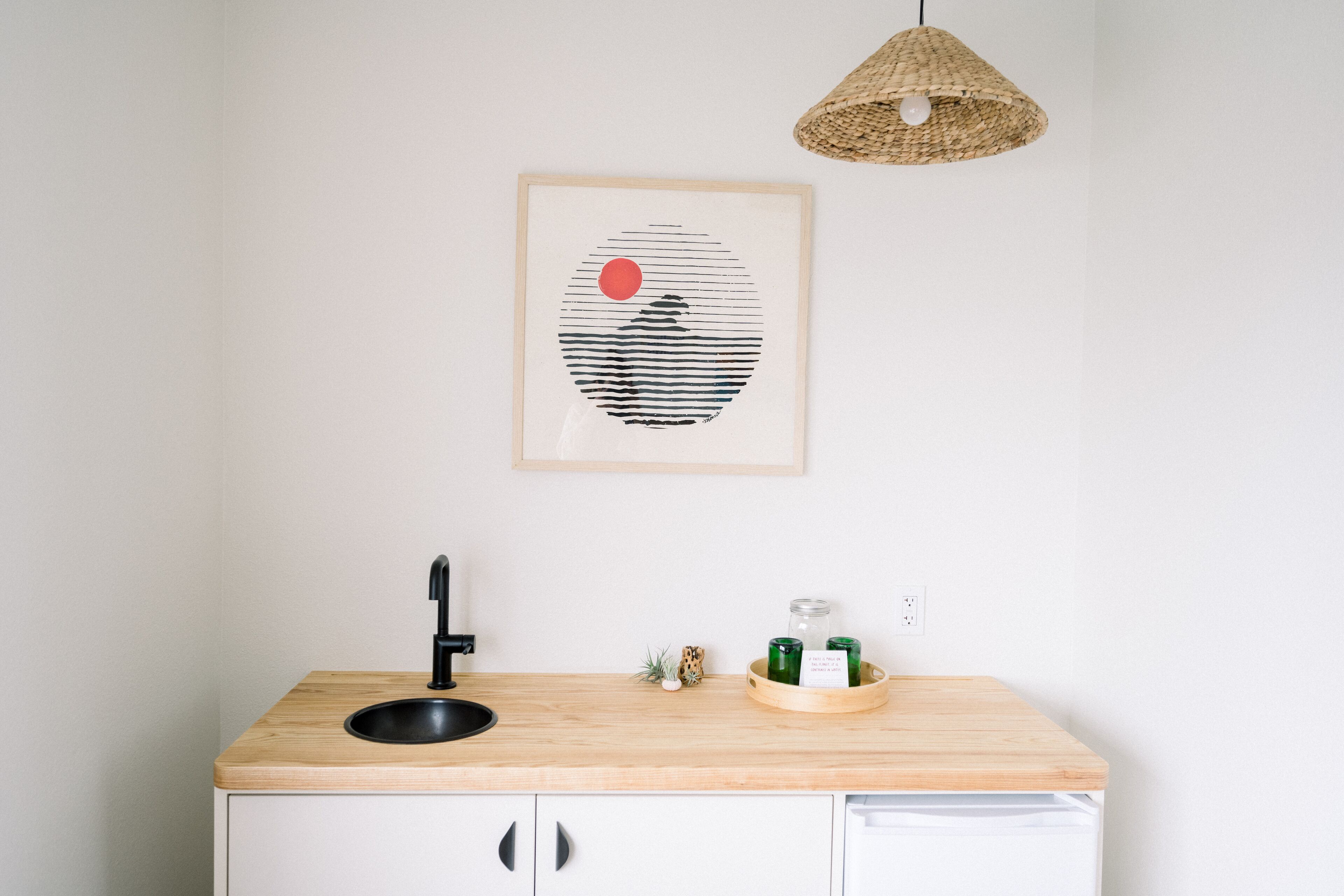 A kitchen with a wooden countertop, a black sink, a woven pendant light, and a framed artwork on the wall.