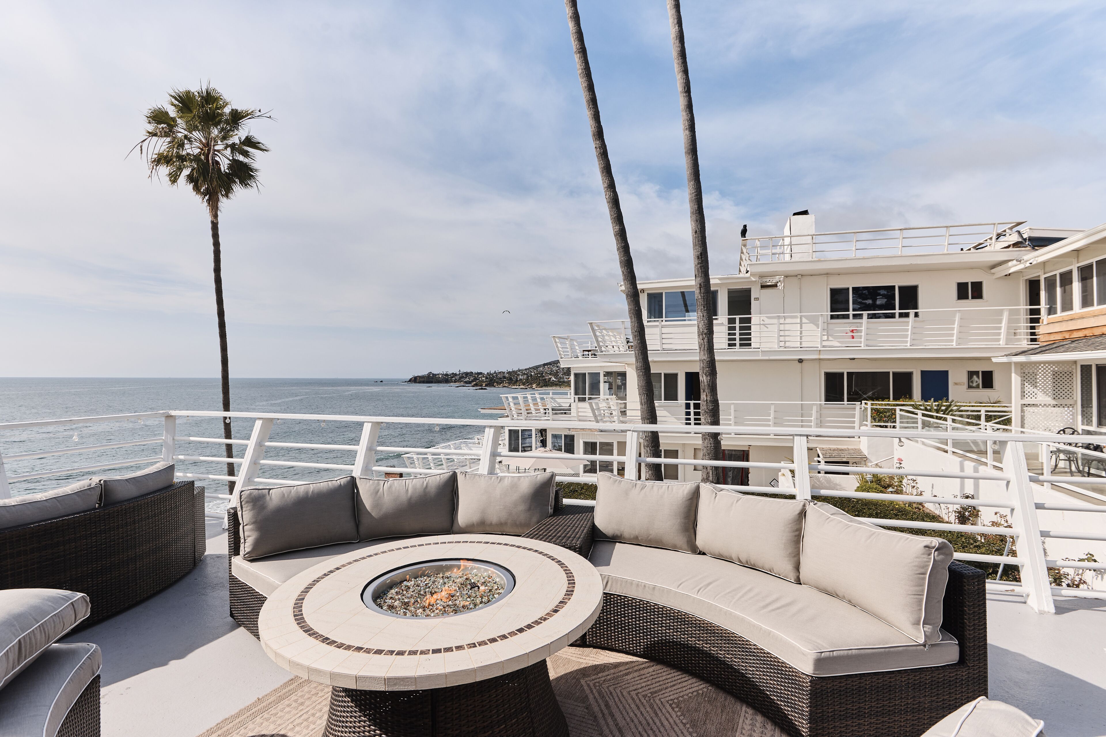 A sunlit outdoor seating area with a round table and cushions, overlooking a body of water and a white building with multiple balconies.