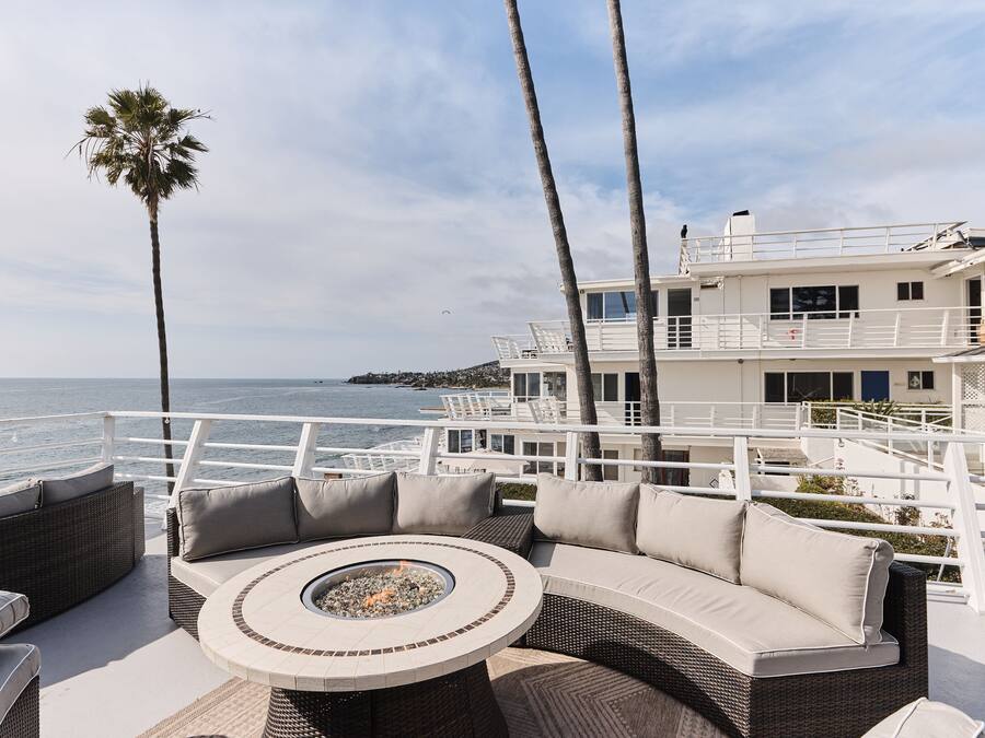 A sunlit outdoor seating area with a round table and cushions, overlooking a body of water and a white building with multiple balconies.