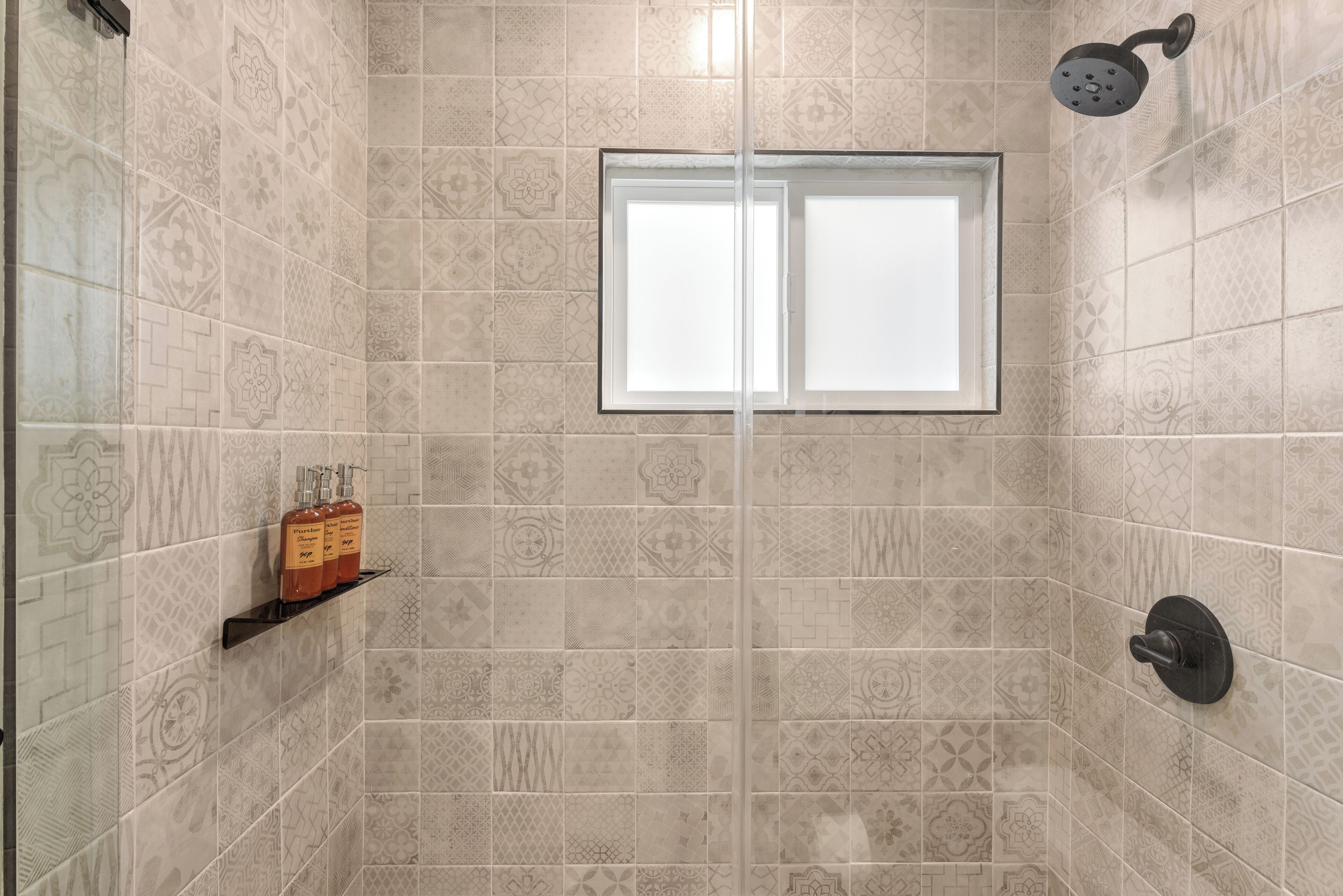 A shower with a window, patterned tiles, and a shelf with bottles.