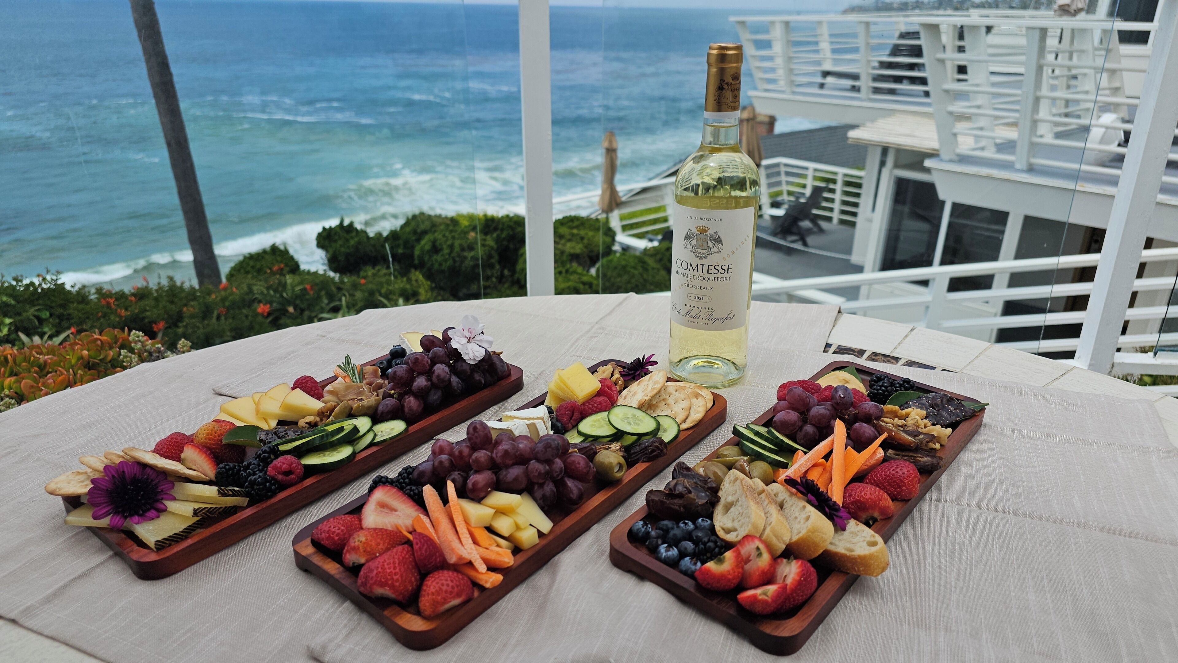 Une table garnie d’une variété de fruits et de fromages, une bouteille de vin et une vue sur l’océan.