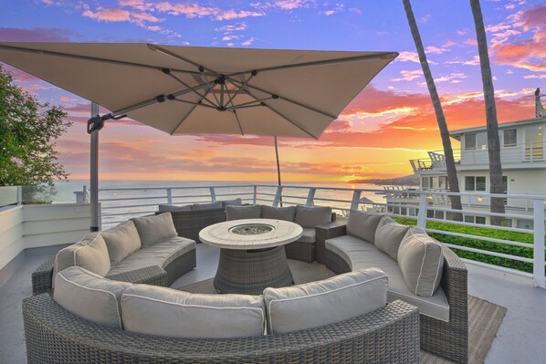 A wicker outdoor seating area with a round table and a large umbrella, overlooking a scenic sunset view.