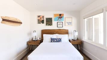 Un dormitorio ordenado con una cama grande, dos mesitas de noche con lámparas y decoración en la pared que muestra escenas de surf y motivos tropicales.
