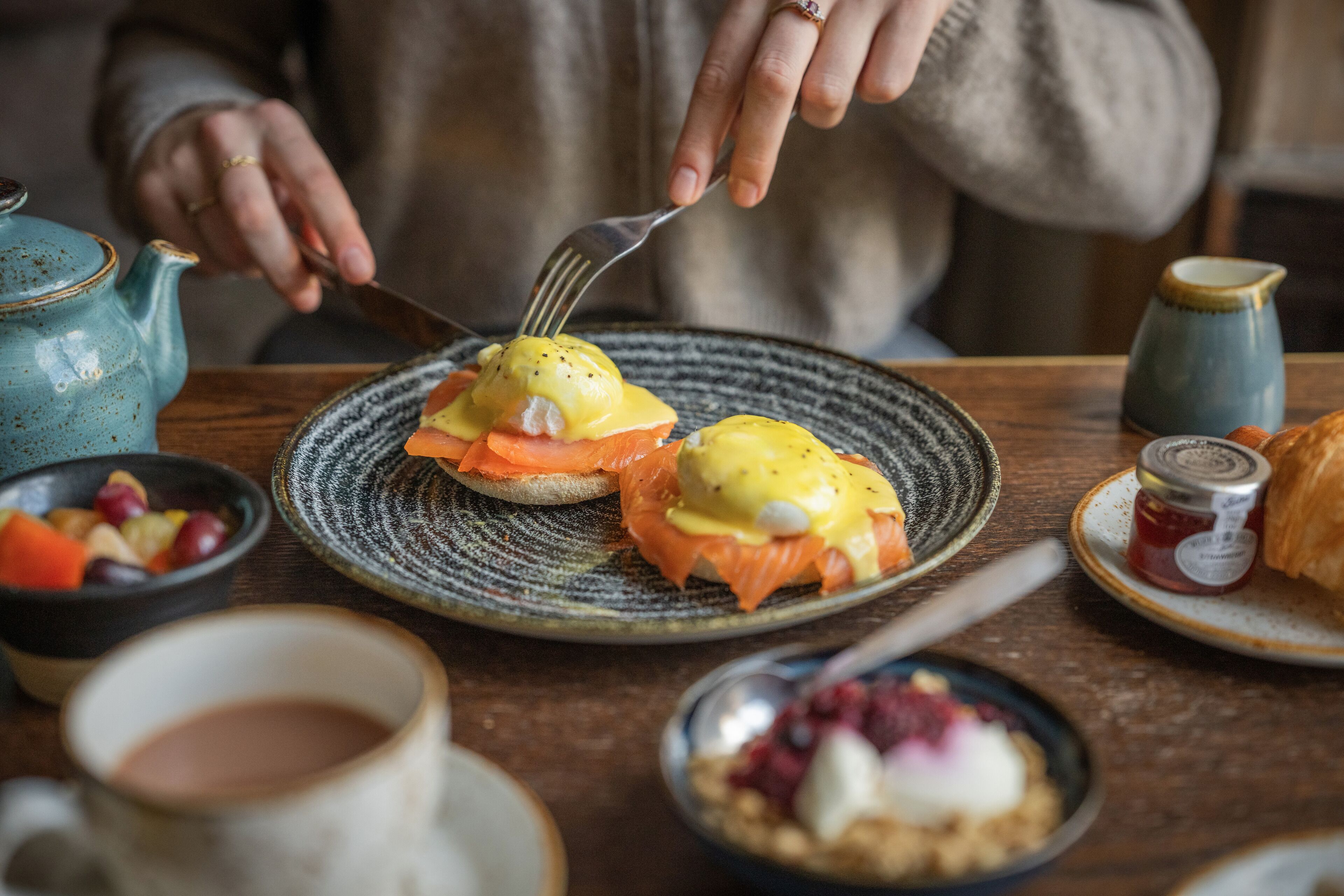 A dining table with a plate of eggs benedict, a teapot, a cup, and a jar of jam.
