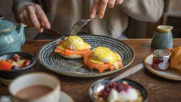 A dining table with a plate of eggs benedict, a teapot, a cup, and a jar of jam.
