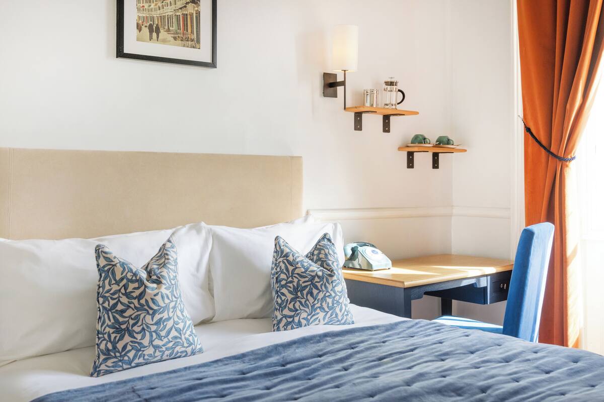 A hotel room with a bed, a desk, a chair, a telephone, and a wall-mounted shelf with a lamp and a mug.