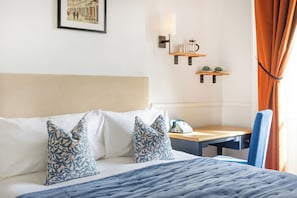 Una habitación de hotel con cama, escritorio, silla, teléfono y estante en la pared con lámpara y taza.