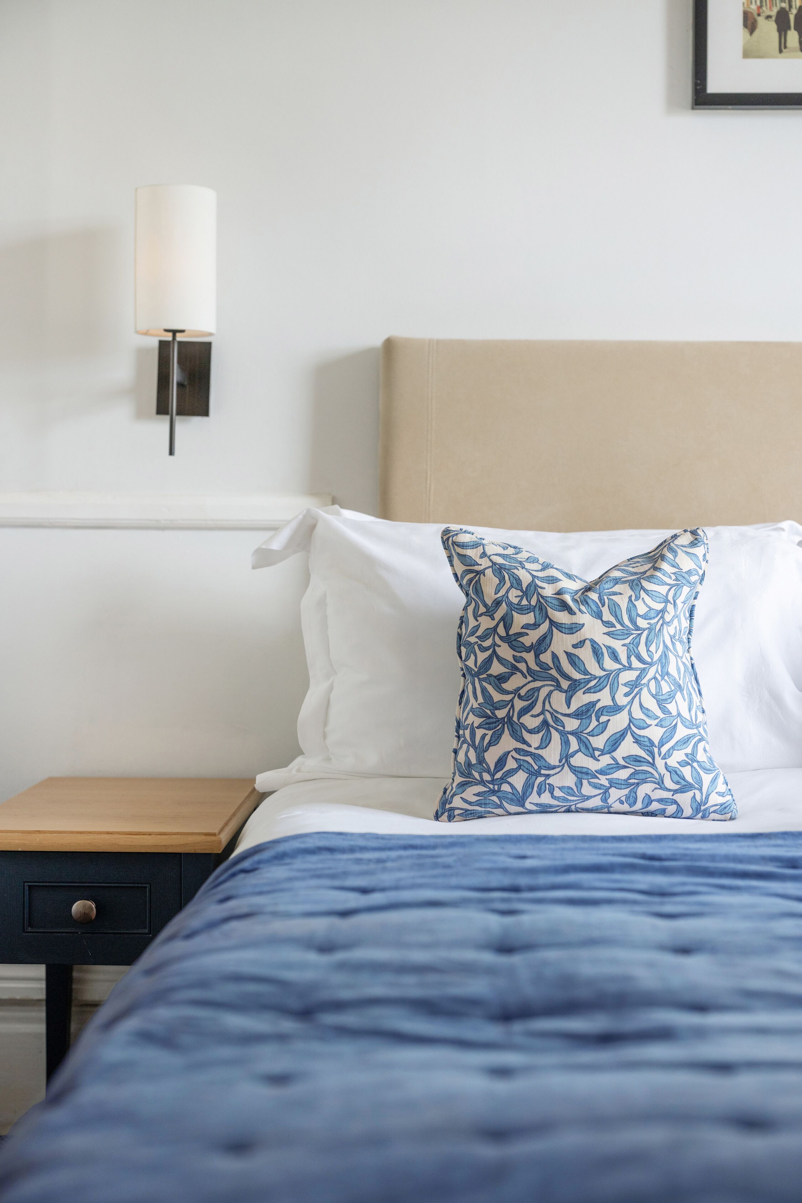 Una cama bien hecha con una colcha azul y una almohada estampada, una mesita de noche con una lámpara, y un aplique de pared.
