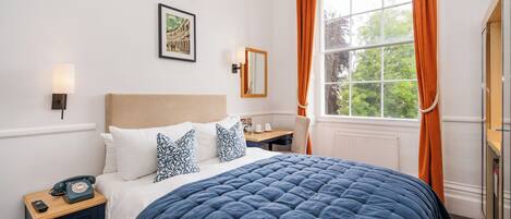 A neatly arranged bedroom with a large bed, a bedside table with a telephone, a desk, and a window with curtains.