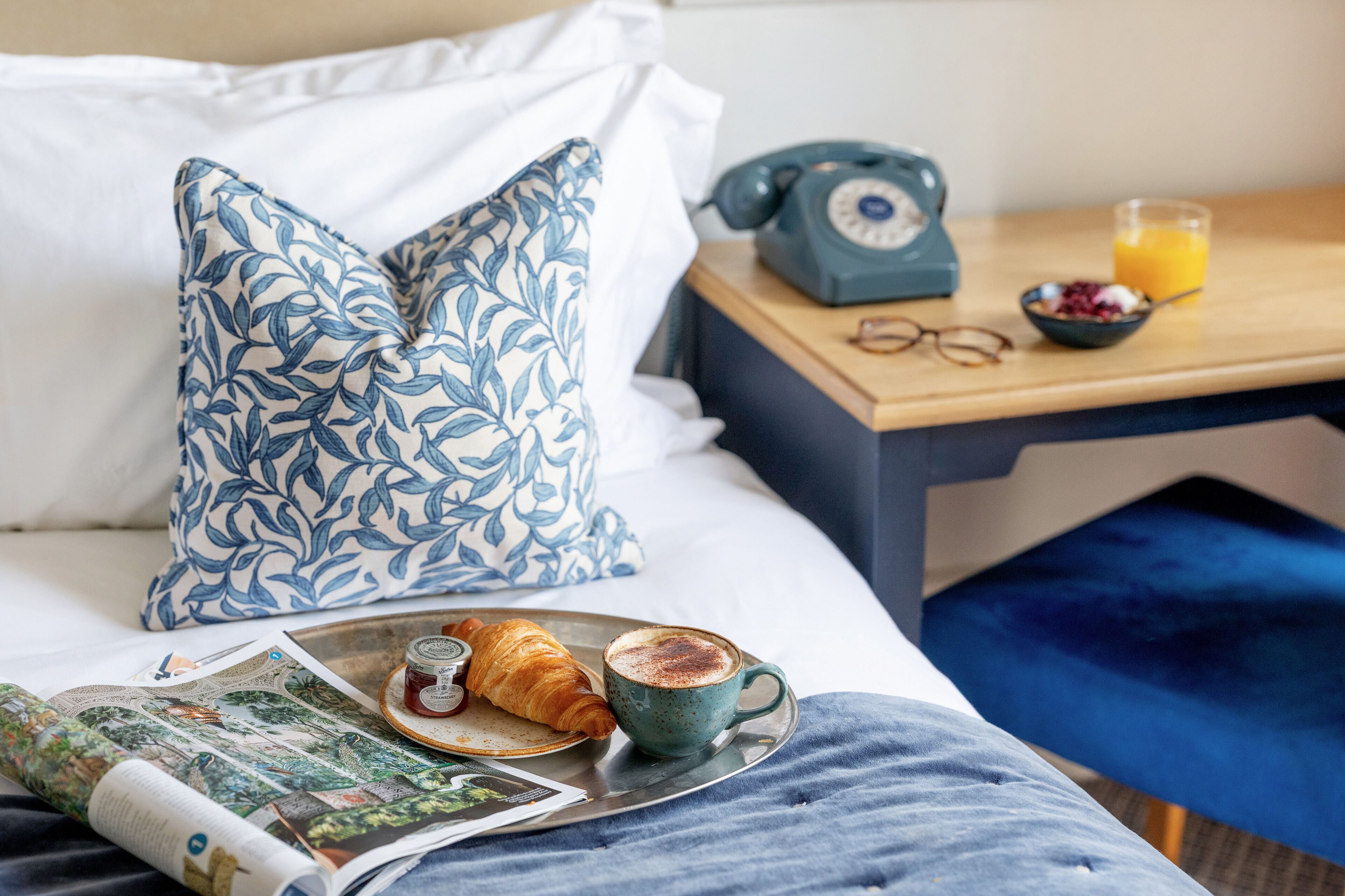 A neatly made bed with a pillow, a bedside table with a rotary phone, a bowl of fruit, and a cup of coffee.