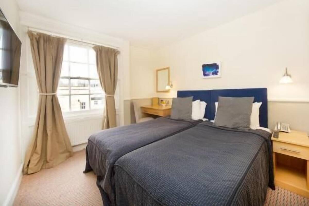 A hotel room with a double bed, a bedside table, a lamp, a window with curtains, and a wall-mounted TV.