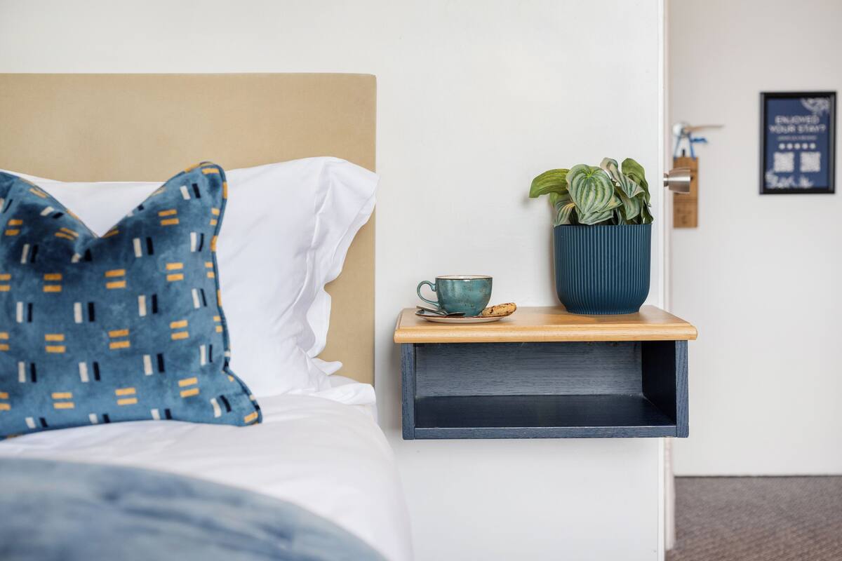 A neatly made bed with a pillow, a bedside table with a cup and a plant, and a wall-mounted sign.