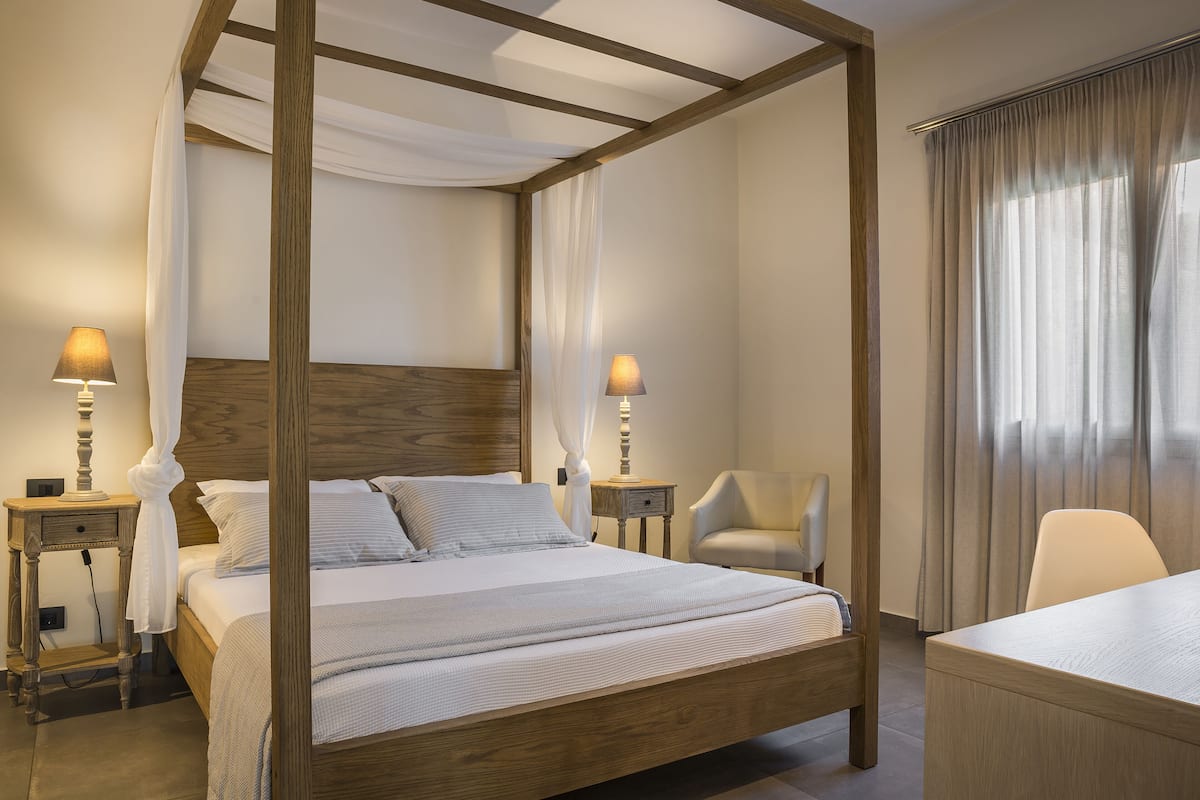 Ein Hotelzimmer mit einem Himmelbett, einem Holzkopfteil, zwei Nachttischen mit Lampen, einem Sessel und einem Schreibtisch.