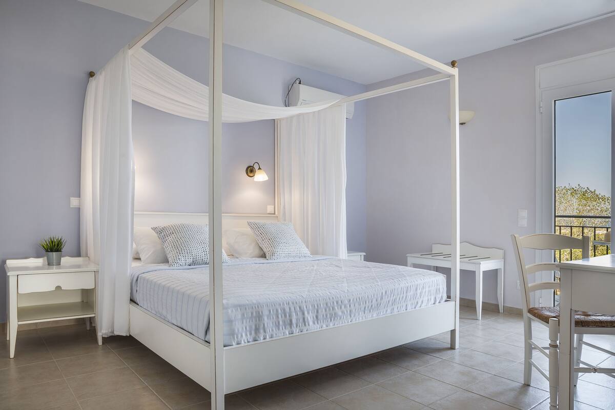 Ein Schlafzimmer mit einem Himmelbett, einem Nachttisch mit Lampe und Blick ins Freie durch ein Fenster.