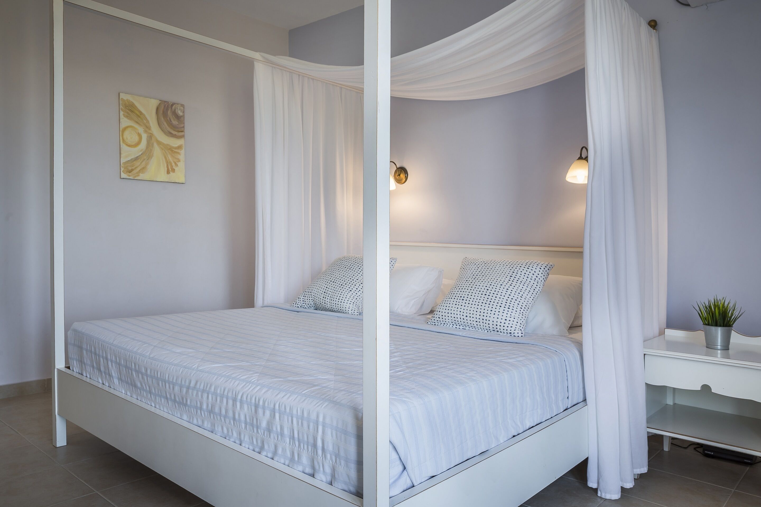 Een hemelbed met wit beddengoed, een nachtkastje met een lamp en een aan de muur hangend kunstwerk.